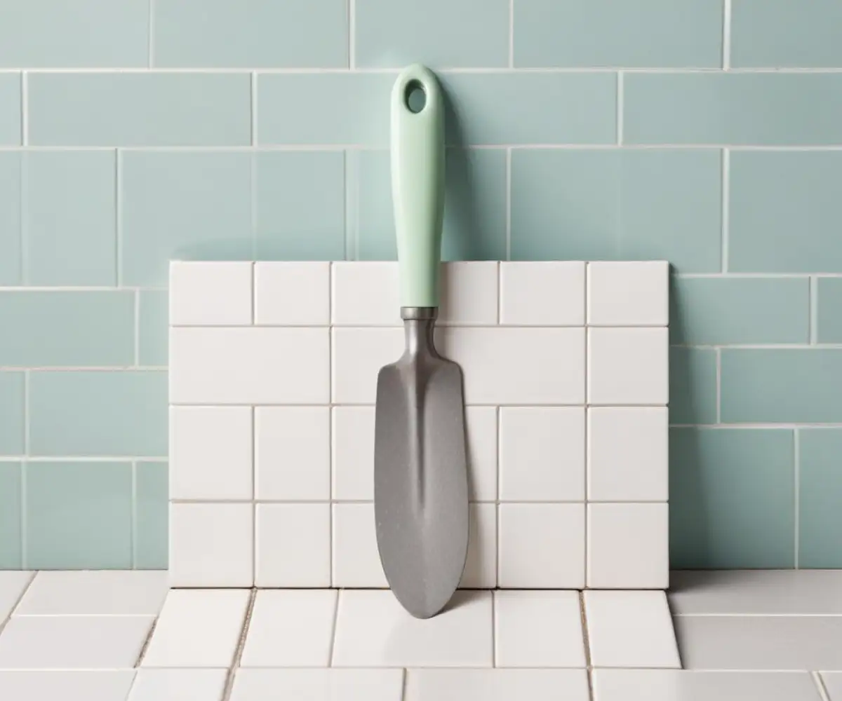 A metal notched trowel resting on a stack of 3x12 inch white ceramic subway tiles.