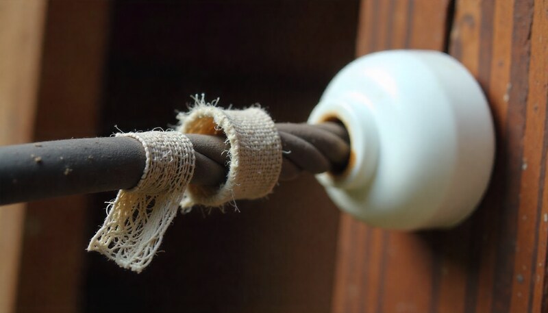 Close-up of aged, cracked, and frayed cloth-covered electrical wires adjacent to a white ceramic knob insulator mounted on a wooden beam.