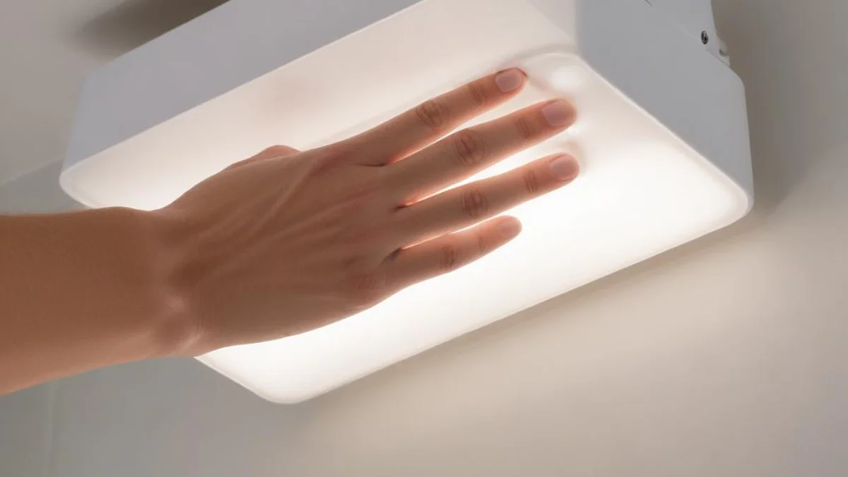 A close-up of a white, rectangular emergency light fixture, with one hand gently prying off the translucent plastic cover to reveal the bulbs inside.