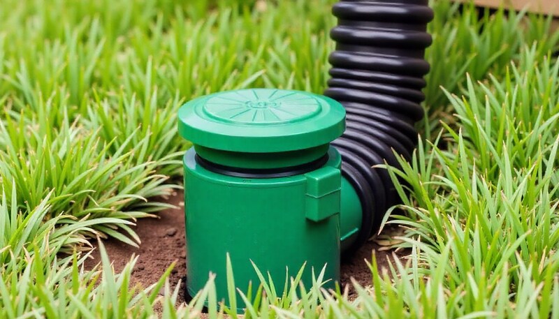 Green pop-up drainage emitter installed flush with a lawn, connected to a black corrugated sump pump discharge pipe.